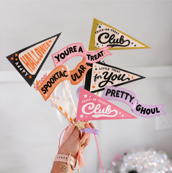 A hand holds a bundle of colorful triangular Halloween-themed pennant flags with playful text like 'Halloween Club,' 'You're a Treat for You,' 'Pretty Ghoulish,' and 'Spooktacular Clue,' against a soft light gray background.